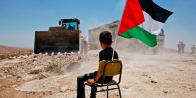 TOPSHOT - A Palestinian boy sits on a chair with a national flag as Israeli authorities demolish a school site in the village of Yatta, south of the West Bank city of Hebron and to be relocated in another area, on July 11 2018. (Photo by HAZEM BADER / AFP)        (Photo credit should read HAZEM BADER/AFP via Getty Images)